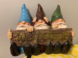 Three gnomes sitting behind a sign reading "Welcome" -- the gnome on the left is wearing a blue hat and is holding the sign, the gnome in the middle is wearing a red hat and is adjusting his hat, and the gnome on the right is wearing a green hat and holding the sign. 